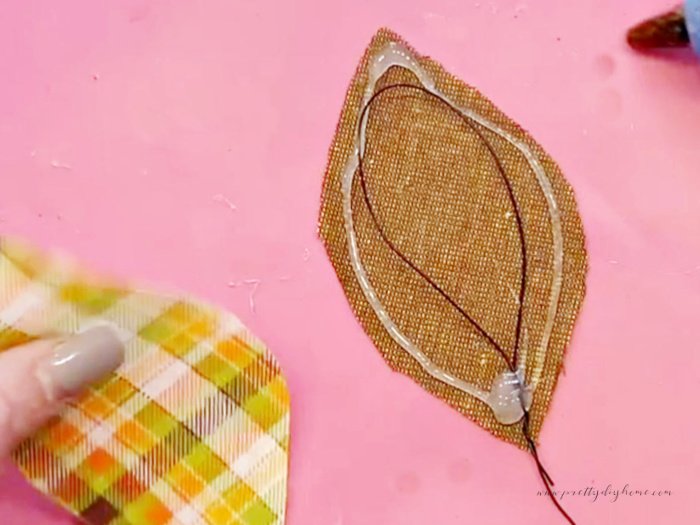 Two pieces of fabric for a fall flower leaf.  The back is brown with a piece of florist wire looped on it.  It is encircled with hot glue.  The second piece of cut plaid fabric is being placed on top to sandwich the wire with the hot glue.