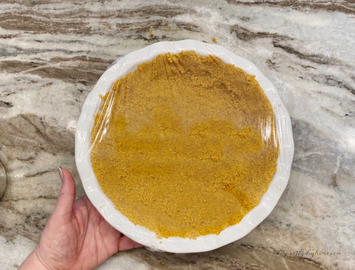 A large porcelain pie plate filld with a fresh graham cracker crust. The plate is wrapped in plastic before being placed in the fridge to cool.