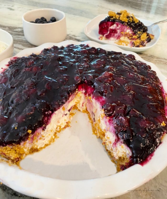 a large blueberry cream cheese pie is cut open wih a piece of pie missing so you can see the layers of graham cracker crust, a thick layer of cream cheese filling and a cooked fresh blueberry topping.