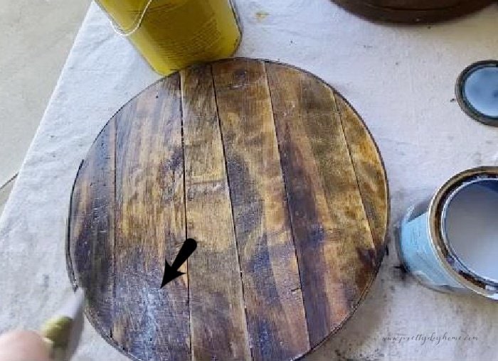 Adding varathane to the outside of a vintage wooden cheesebox with an arrow showing the part of the box with rough wood that needs extra attention and varathane thinning.