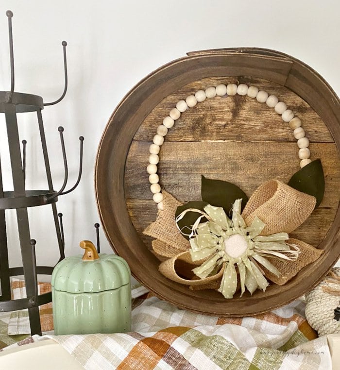 A restored vintage wood cheesebox sitting on top of a cabinet. The cheesebox is framing a simple sunflower wreath and its surrounded by small pumpkins.