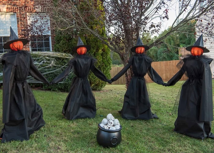 Four large DIY outdoor Halloween witch decorations holding hands around a cauldron of skulls. Each with has an orange pumpkin face, black hat and pretty tulle capes.