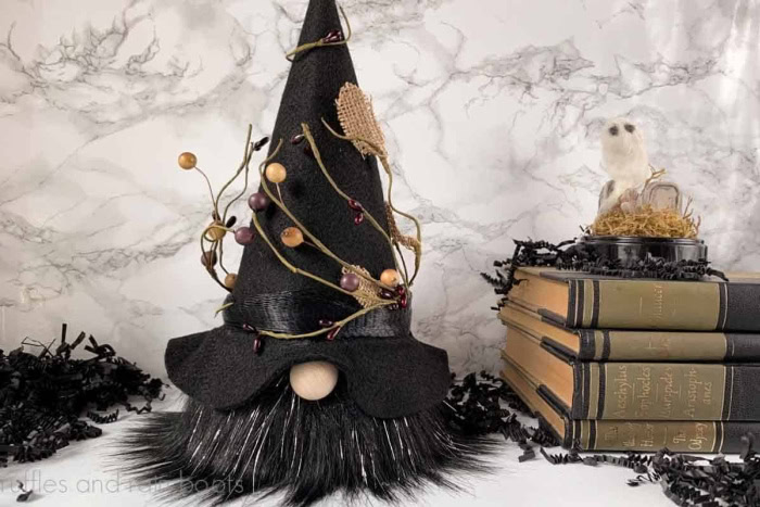 A fun DIY witch gnome with black whiskers and a felt witch hat. The hat is decorated with branches and beads, and there is a stack of old books with a small owl sitting beside it.