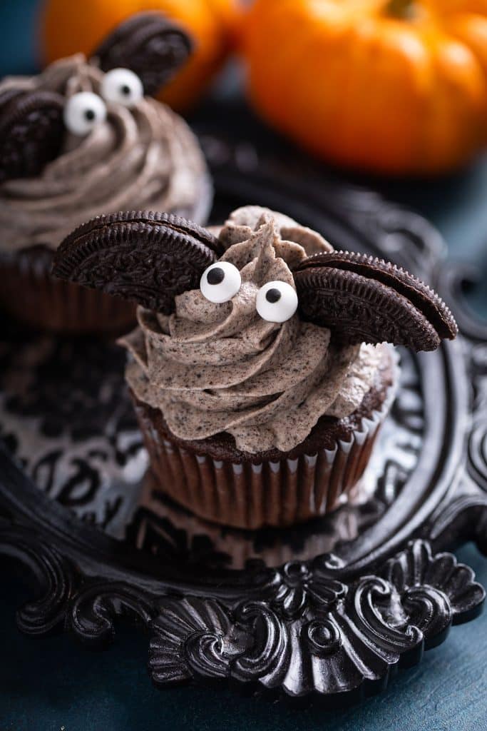 A Halloween cupcake made with dark chocolate then decorated with whipped chocolate icing two candy eyes and a split oreo cookie. The candy eyes and oreos look like a bat sitting on top of the cupcake.