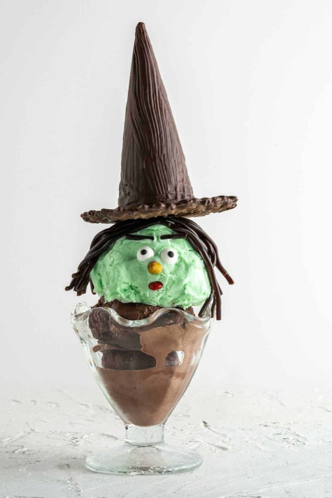 A witch themed chocolate and mint ice cream sundae. The top scoop is green with candy eyesballs and licorice eyebrows and hair. Then its topped with a large cookie witch hat.