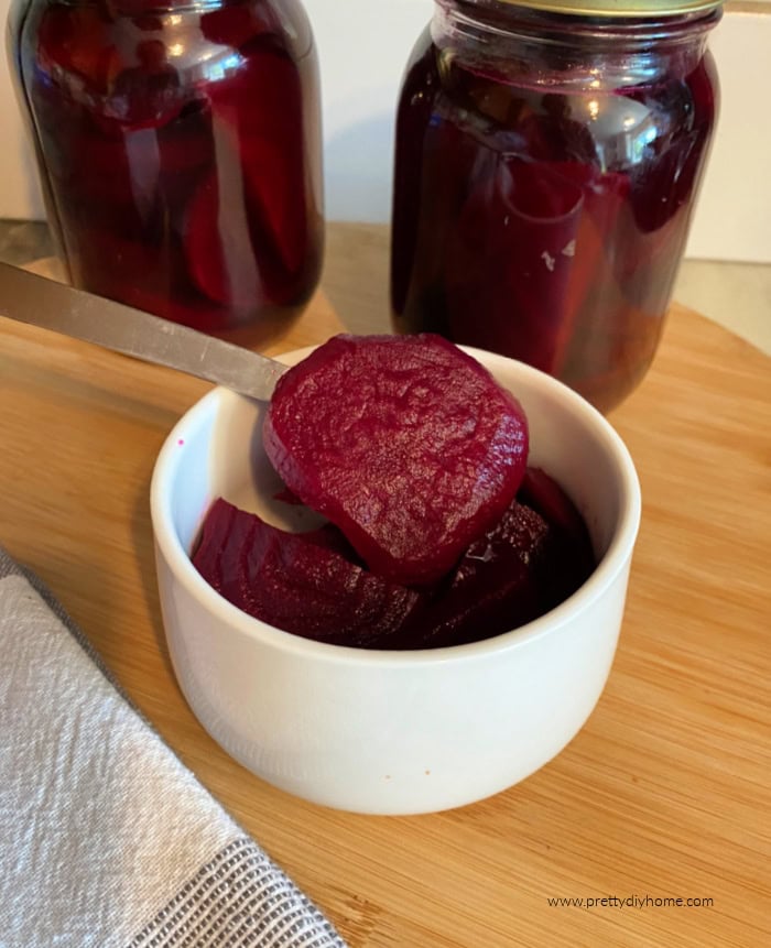 Canned pickled beets being served in a small white bowl. The white bowl is sitting on a bamboo chopping board with a checkers teacloth on the side. You can see two pint sized jars of the spicy pickled beet recipe in the background.