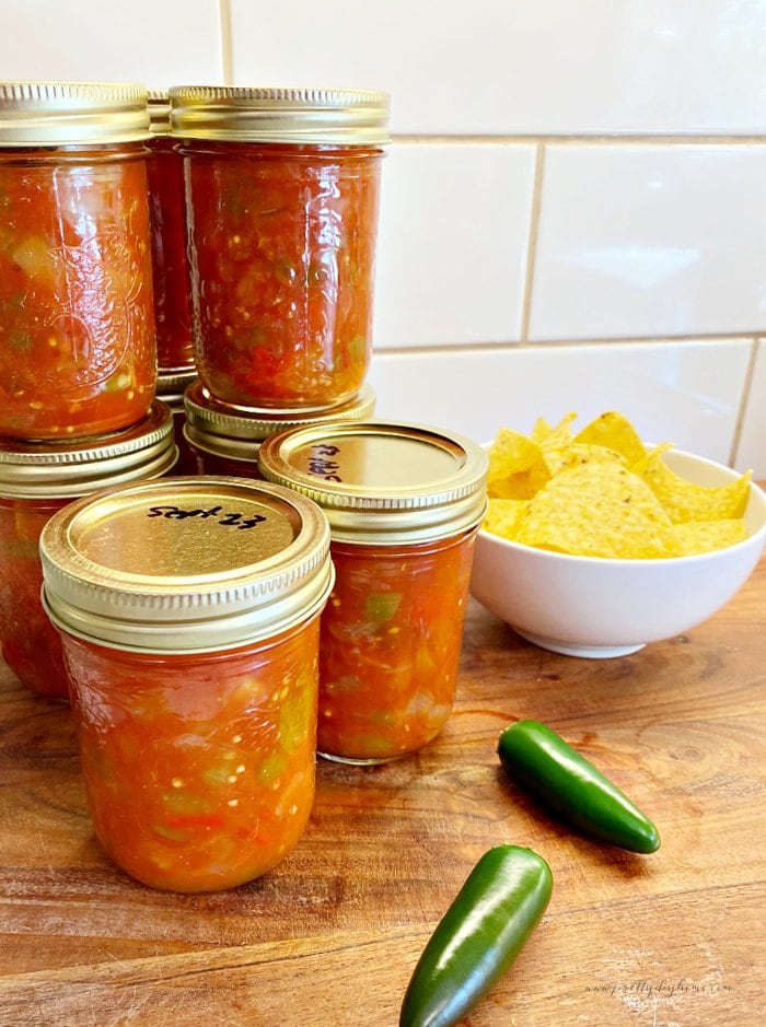 A stack of jars filled with a homemade chunky tomato salsa recipe. The canning jars of salsa are sitting on a wood cutting board with a small bowl of tortilla chips and two green jalapenos.