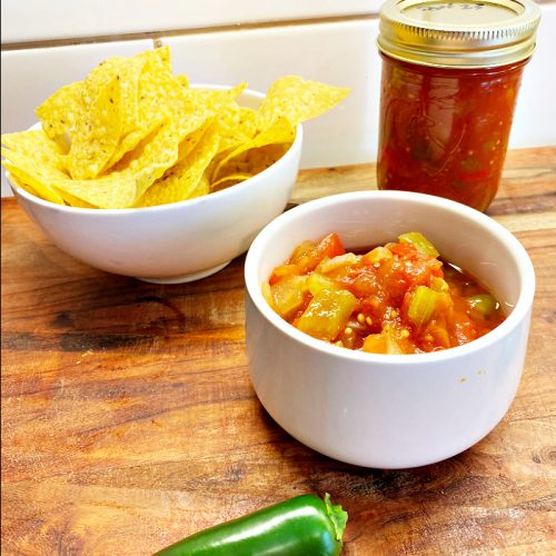 Homemade chunky salsa sitting in a small white bowl , with a fresh jalapeno on the cutting board, a small bowl of tortilla chips, and a jar of freshly canned tomato salsa.