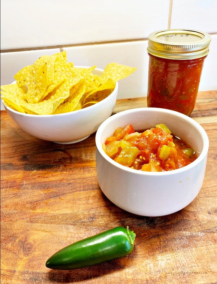 Homemade chunky salsa sitting in a small white bowl , with a fresh jalapeno on the cutting board, a small bowl of tortilla chips, and a jar of freshly canned tomato salsa.