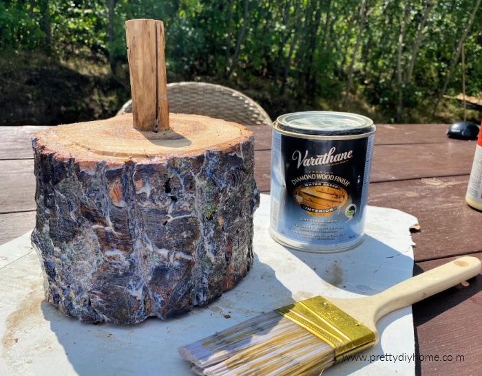 A log pumpkin covered with a coat of fresh varathane.  The can of varathane and the paintbrush are sitting beside the pumpkin on an outdoor table.