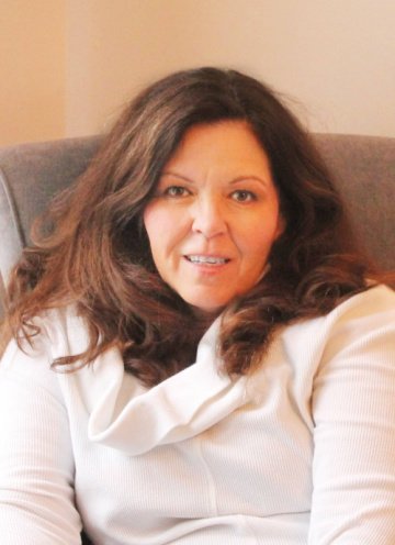 Image of the author Leanna sitting in a chair with long brown hair and a white shirt.