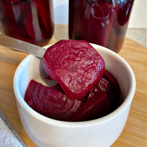 Freshly made spicy pickled beets being served in a small white bowl. One of the sliced beets is being held upto show off the deep burgundy color of the beets.