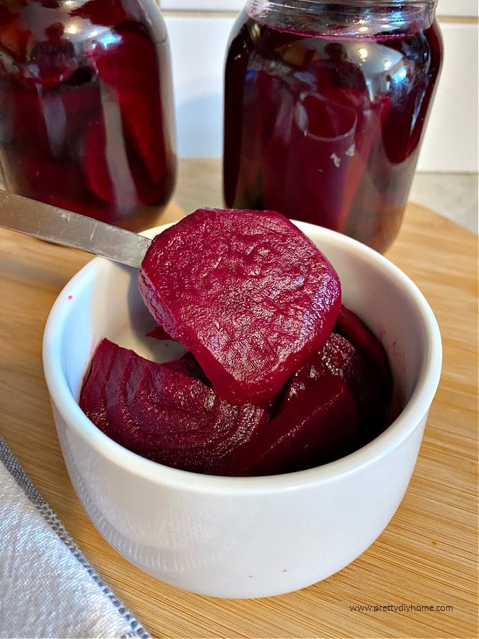 Freshly made spicy pickled beets being served in a small white bowl. One of the sliced beets is being held upto show off the deep burgundy color of the beets.