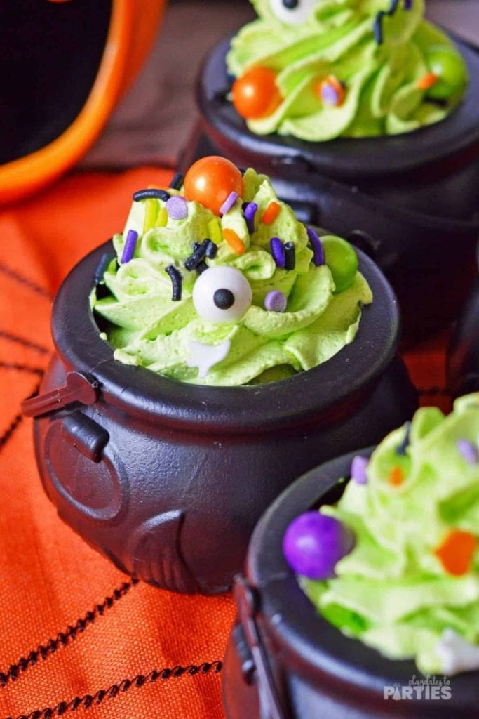 Little black plastic witch cauldrons filled with individual servings of pudding and then topped with colorful purple and green swirls of icing with Halloween candy sprinkles.