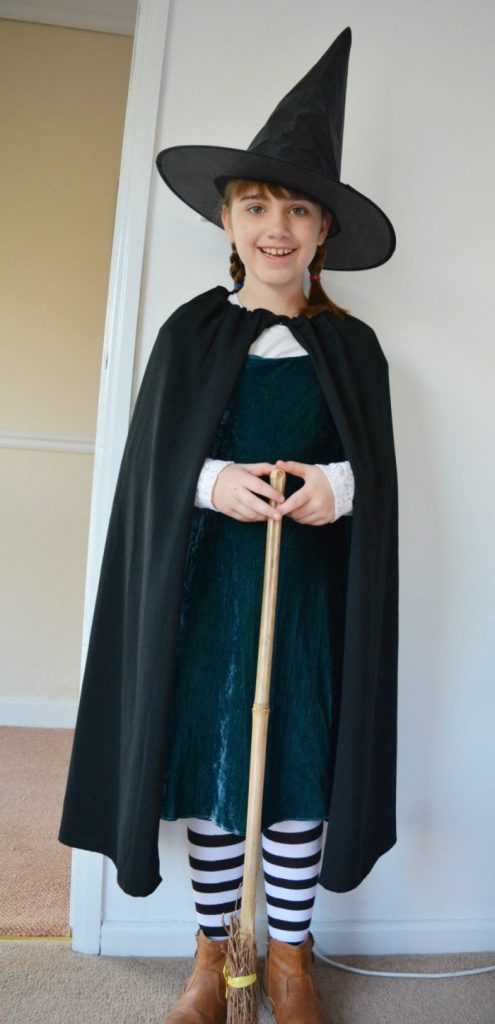 A smiling little girl in a DIY witch costume. She is holding a witch broom and has black striped stockings.