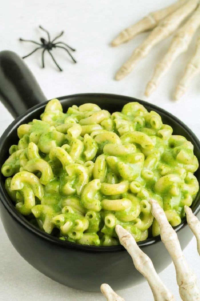 A black bowl filled with bright green macaroni. There is a skeleton hand against the bowl,and a plastic spider in the background.