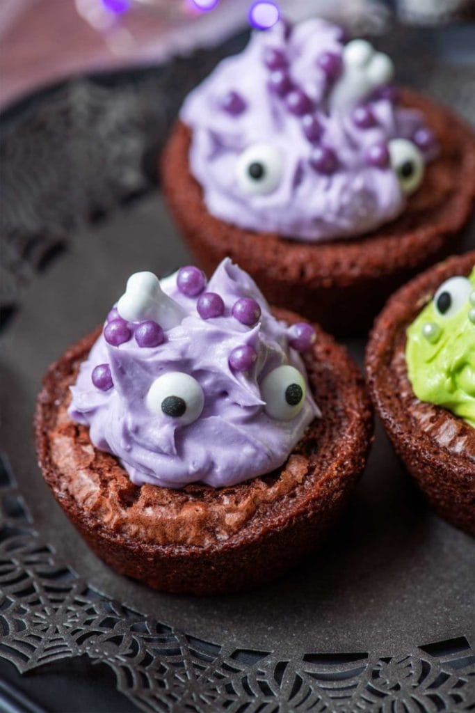 Small round brownies covered with large swirls of icing, some icing is green some is purple. The brownies are then topped with adorable candy eyeballs and different colored Halloween sprinkles.