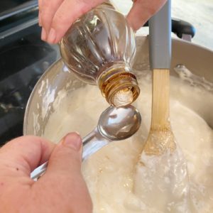 Adding a teaspoon of vanilla extract to a saucepan of melted marshmallow mixture while making a popcorn ball recipe.