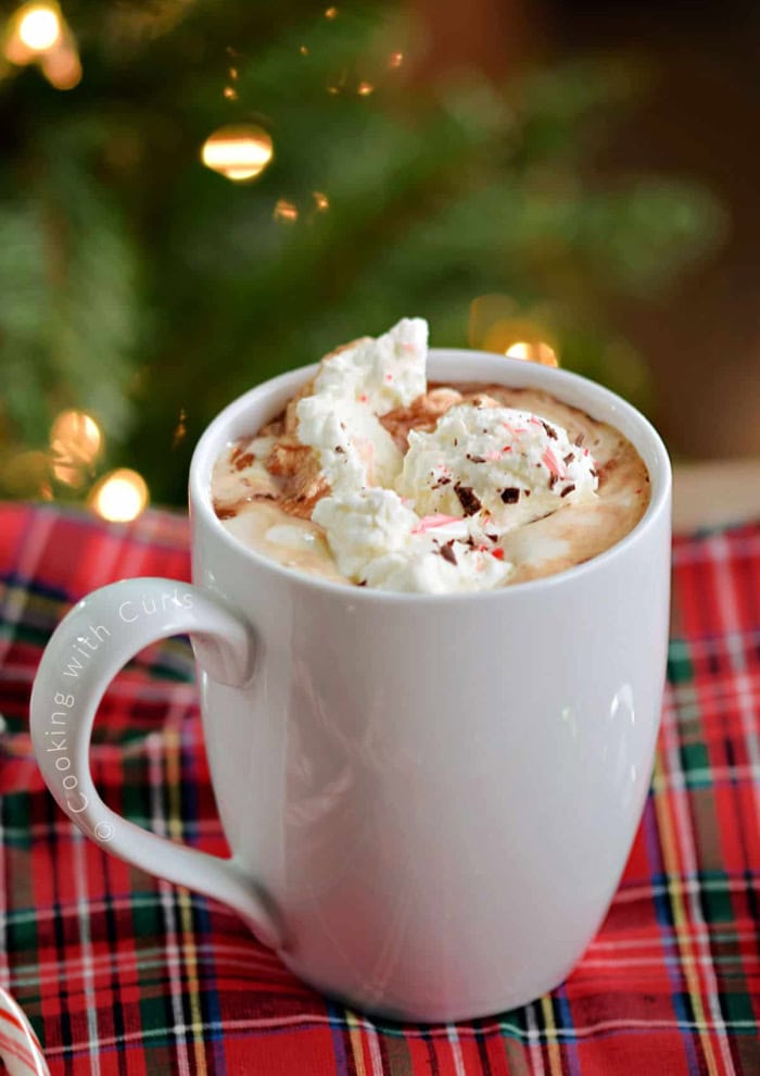 A white mug filled with boozy hot chocolate made with Bailey's then topped with a large swirl of whipped cream and Christmas sprinkles.  The mug is sitting on a red plaid cloth and a Christmas tree is in the background.