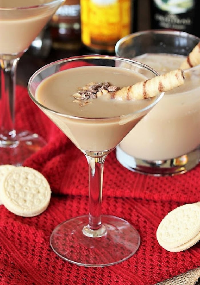 A martini glass filled with a buttered tofee cocktail for Christmas, Its served with a cookie stir stick, and surrounded by Christmas tablecloth and other treats.