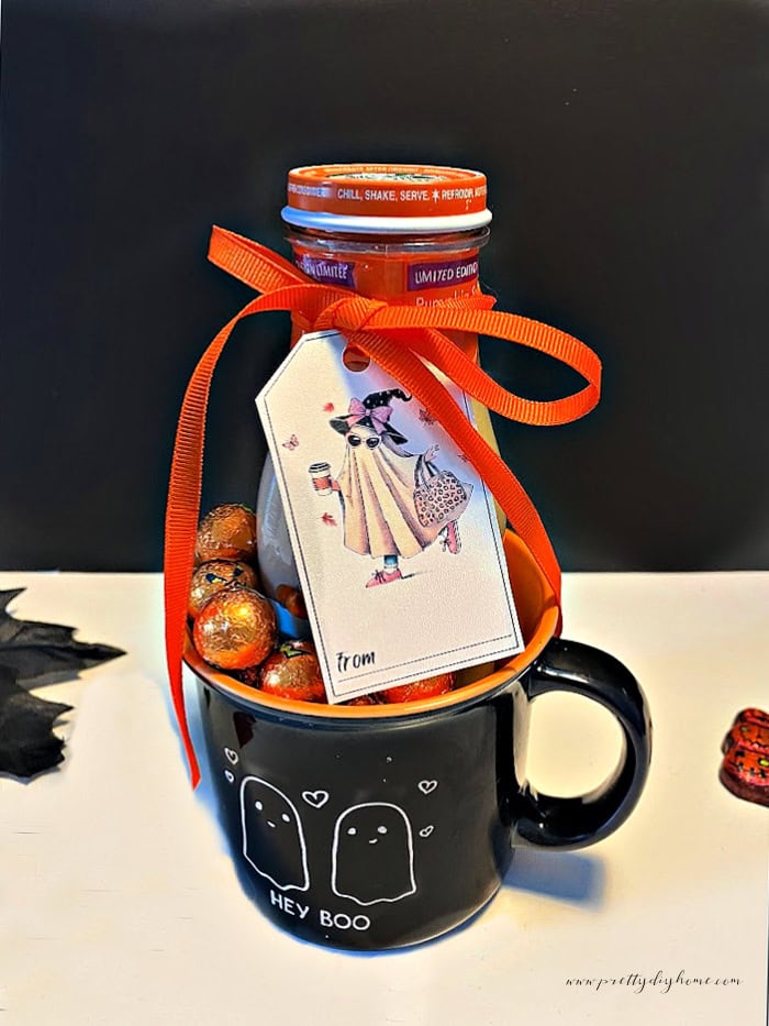 A Halloween mug with a jar of Starbucks iced coffee and chocolate pumpkin inside.  The Halloween gift has a ghost holding coffee themed free printable Halloween tags attached to it