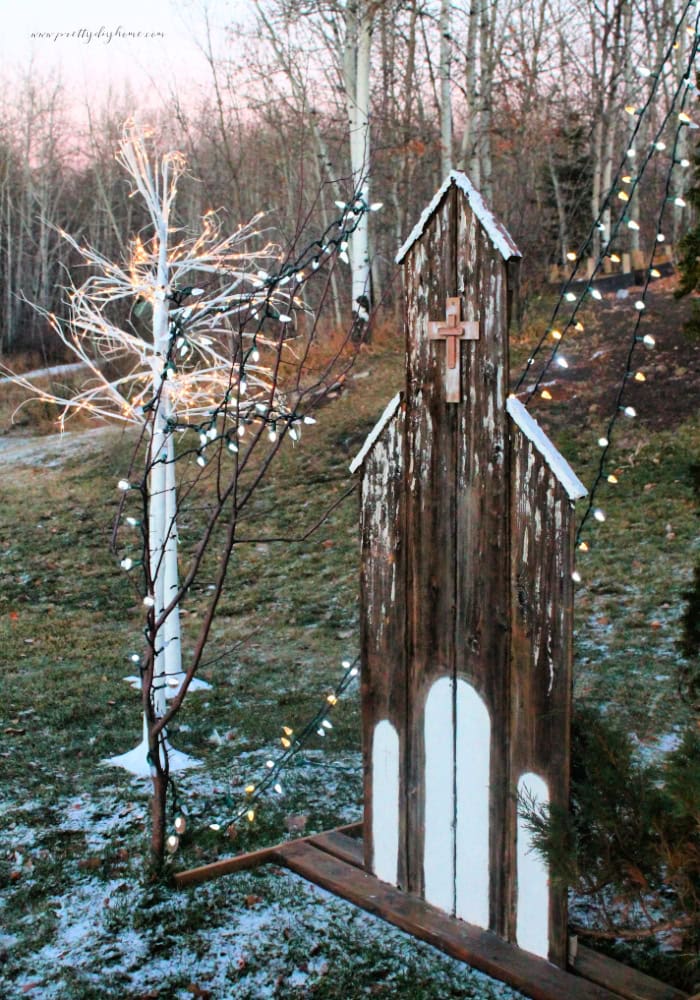 A gorgeous Christmas yard decorated with a DIY rustic church made with old scrap wood. The church is surrounded with white sparkly lights and artificial white birch trees.