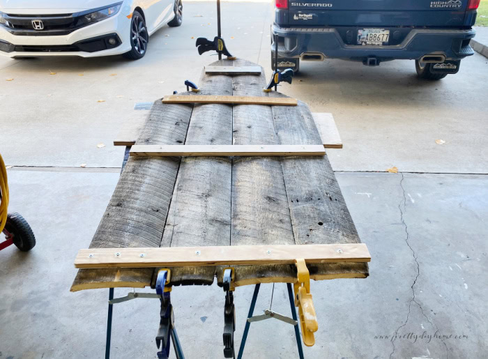 The backside of a DIY outdoor church Christmas decoration showing four slats of old barn wood thats been glued and held together withfour cross pieces of scrap lumber.  It also shows all the clamp placement.