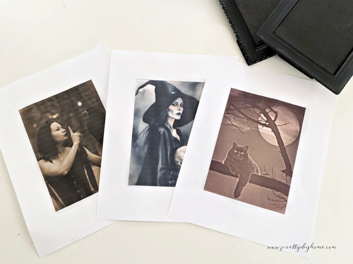 Three different Halloween printables lined up on the counter before placing in frames. There is a witch with a raven, a beautiful witch in a gorgeous black witch hat and a black cat sitting in front of the full moon.