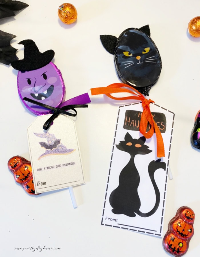 Free printable halloween gift tags attached to chocolate candy suckers the witch one candy has a witch themed halloween tag, and a black cat one has a cat themed halloween tag that says Happy Hauntings. The back ground is white with pumpkin candies scattered on to it.