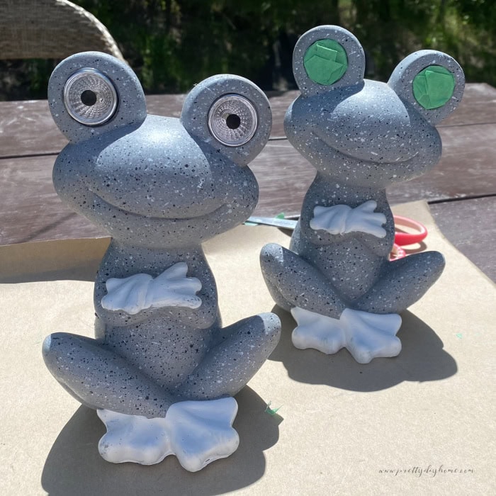 Two grey outdoor garden frogs, one has it eyes covered in painters tape in preparation of getting painted into a DIY outdoor Halloween solar witch frog.  The solar panel and the light up eyes need to be taped off to protect from paint.