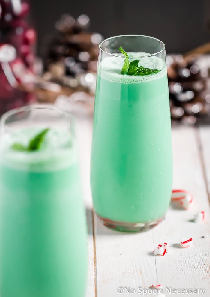 Two tall thin glasses of grinch green cocktail with Baileys.  There are frosted pinecones and red berries in the background.