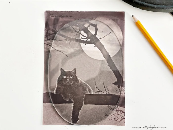 Placing an oval glass frame front onto an black cat Halloween picture. There is a pencil beside to trace the image for cutting for a Halloween frame diy projec.t