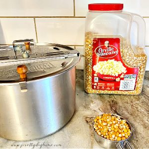 A half cup of popcorn kernels in a measuring cup, a large container of Orville Redenbakers popcorn and a stove top popcorn maker, all sitting on a countertop before starting to make popcorn balls.