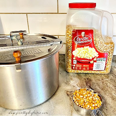 A half cup of popcorn kernels in a measuring cup, a large container of Orville Redenbakers popcorn and a stove top popcorn maker, all sitting on a countertop before starting to make popcorn balls.