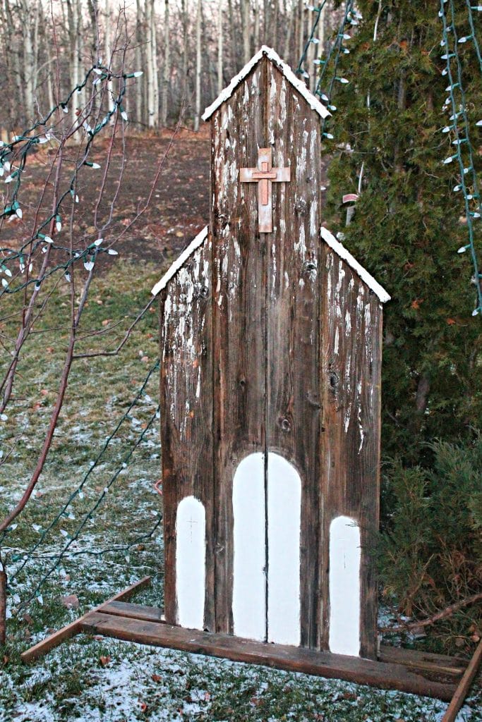 A six foot tall outdoor Christmas decoration made from wood.  The wooden church looks very old with white plaster doors and a metallic cross.