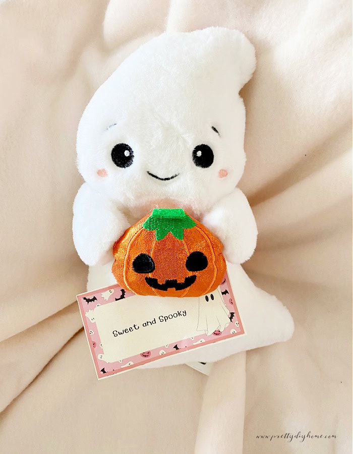 A sweet and spooky ghost free printable Halloween gift tag attached to a sweet stuffed ghost toy. The background is a soft peach colored baby blanket.