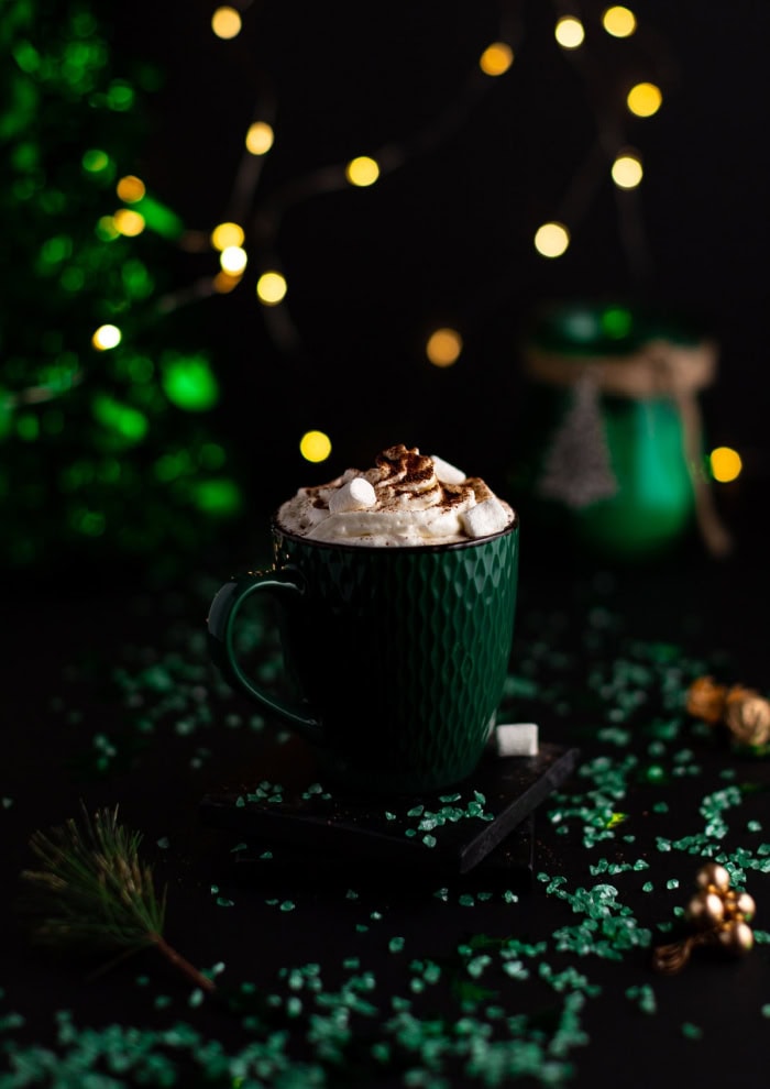 A dark image with a dark green mug filled with Bailey's hot chocolate.  The hot chocolate has a thick whipped cream topping with marshmallows, and in the dark bacground you can see a second mug of hot chocolate and Christmas lights.