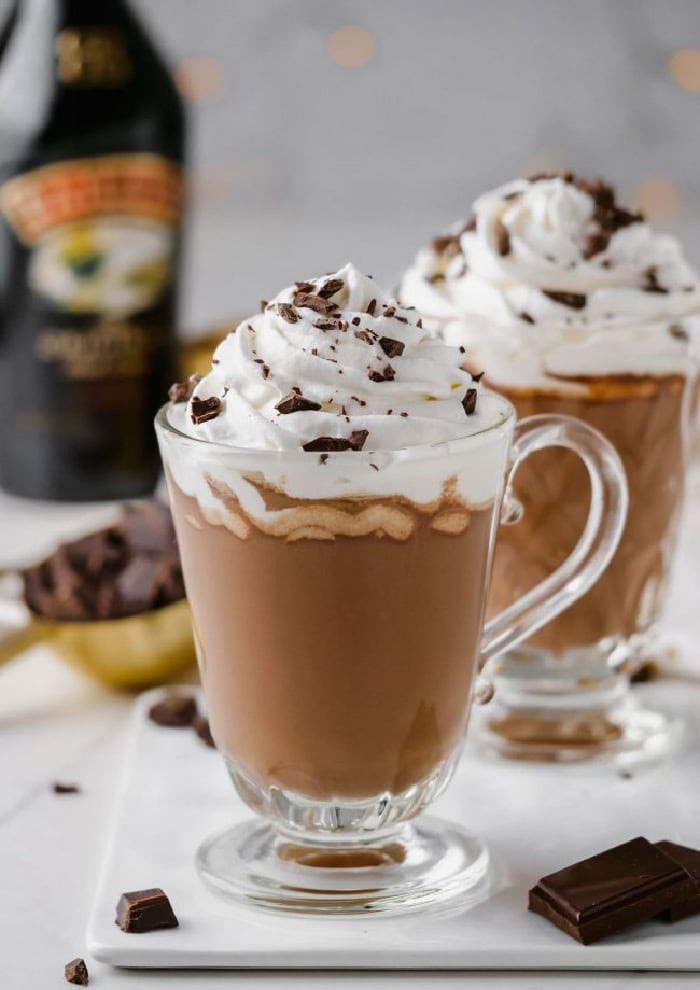 Two tall clear mugs filled with creamy hot Bailey's hot chocolate with large swirls of whipped cream and chocolate bits.  