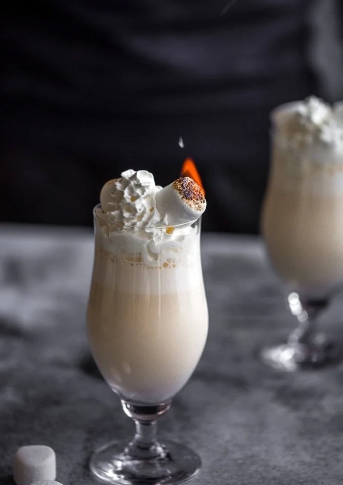 A baileys winter cocktail in a tall class with a toasted marshmallow on top.  The background is dark and cozy looking,