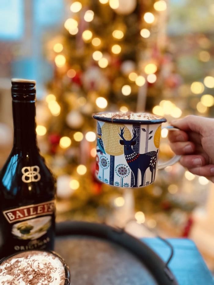 A mug of Bailey's hot chocolate being held by hand in front of a Christmas tree.  You can see whipped cream on top of the hot chocolate and its being served in a blue Christmas mug.