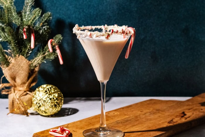 A tall martini  glass filled with a andy cane martini.  The glass is rimmed with crushed candy canes, and there is a mini candycane hanging on the side.