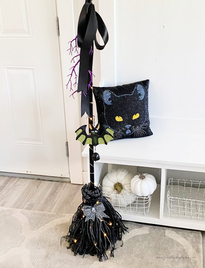 A Dollar Tree witch broom DIY thats mostly black with sparkling accessories, and miniature fairy lights twinkling. There is a black cat pillow beside it along with some white faux pumpkins.