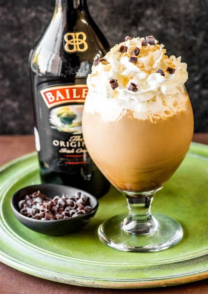 A large stemmed glass filled to overflowing with Irish coffee, then covered with thick whipped cream and chocolate curls on top.  The drink is sitting on a green tray, with a small container of chocolate curls beside it and a bottle of Bailey's in the background.