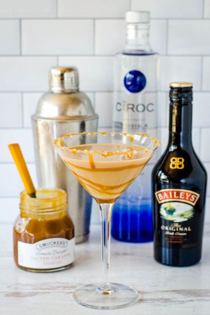 A salted caramel martini in a martini glass, you can see a bottle of Bailey's a jar of salted caramel, a bottle of vodka and a shaker in the background. A beautiful Christmas cocktail with Baileys.