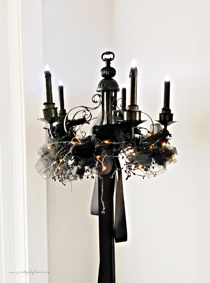 A Halloween DIY Chandelier, DIY scary decoration for the front yard. Its a beautiful chanderlier decorated in all black with a long satin ribbon bow, black feathered crows, fairy lights, beaded garland and black tulle. An easy to make outdoor Halloween prop.