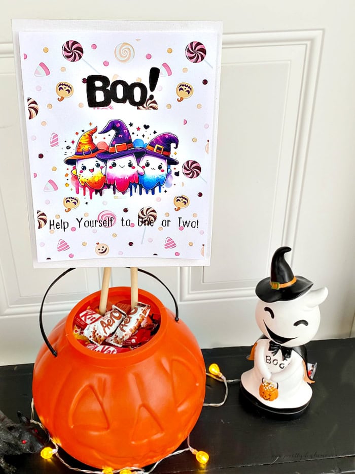 A ghost themed Halloween candy sign decorated in a large plastic pumpkin with a small ghost on the side.  Its sitting on a Halloween porch on top of a black bench, right in front of the door for trick or treaters.