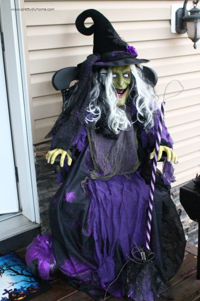 A life sized DIY scary Halloween witch prop sitting on the front porch, she is purple and black sitting on a chair with a green face and a witches broom.