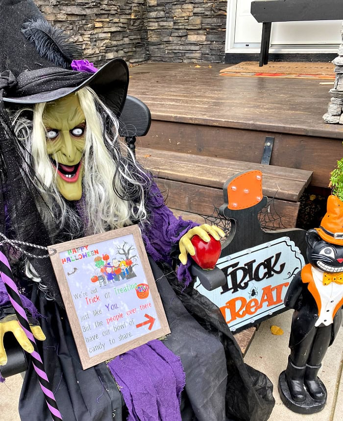 A Halloween porch decorated with a large witch holding a framed Halloween candy sign that directs children next door for candy.