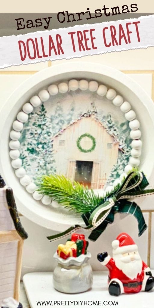 A dollar tree pie plate turned into a Christmas sign using a free printable of a white barn, decorated with white wood beads, greenery and a small green plaid bow.