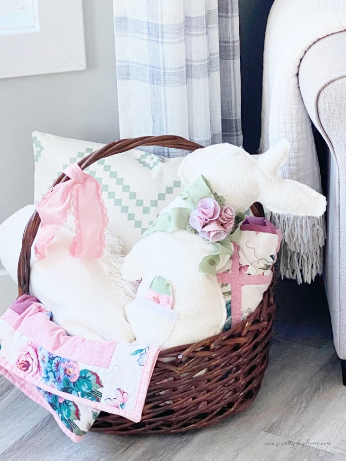 A homemade bunny pillow in a sweet bunny shape. The bunny pillow is cream colored with a soft sage green ribbon, andd homemade pink fabric flowers around the neck. It also has a lace trimmed pocket on the front thats holding a diy fabric Easter egg in green, pink and white.